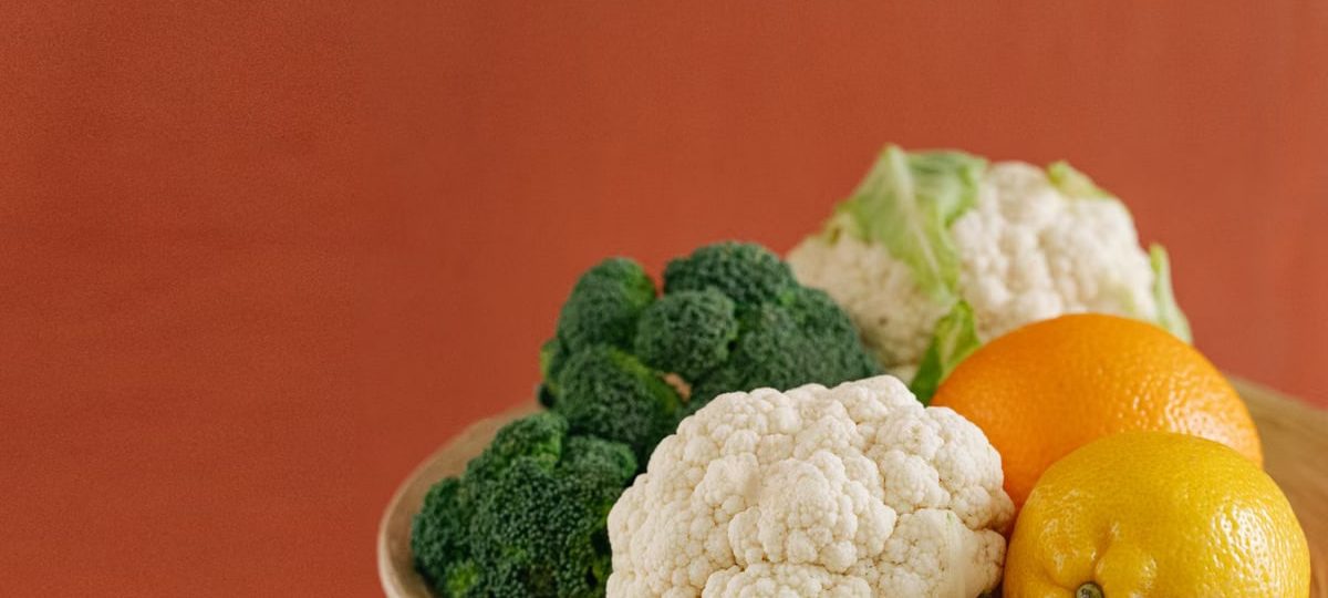 Close-up of fresh broccoli, cauliflower, lemon, and orange on a wooden plate with an earthy background.
